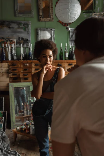 Seductive african american woman looking at blurred man in art studio with vintage mirrors and paintings — Stock Photo