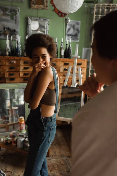 Cheerful african american woman covering mouth while laughing near blurred man in workshop with vintage furniture and mirrors — Stock Photo