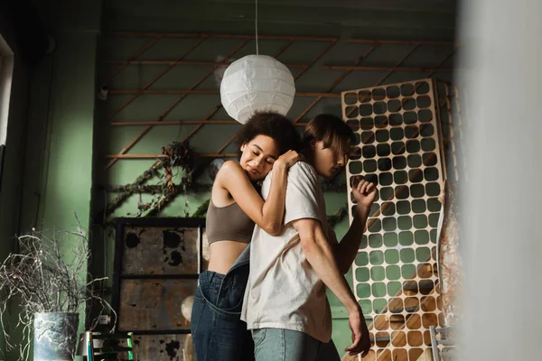 Bonita mujer afroamericana con los ojos cerrados abrazando joven artista en estudio en primer plano borrosa - foto de stock