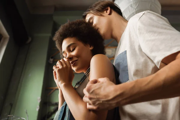 Vista basso angolo di giovane uomo abbracciando sensuale donna africana americana sorridente con gli occhi chiusi in studio d'arte — Foto stock