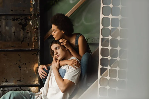 Pretty african american woman sitting on stairs and hugging pensive boyfriend looking away in workshop — Stock Photo