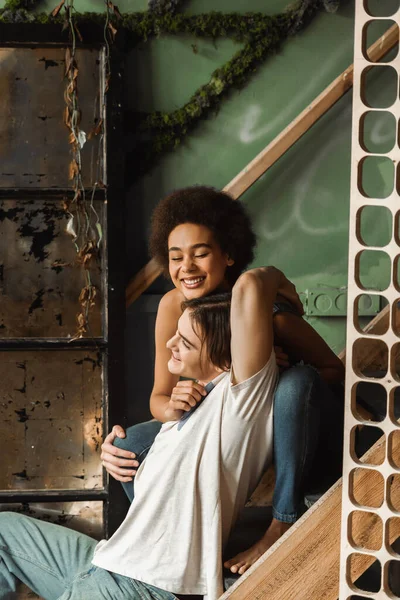 Cheerful african american woman with closed eyes embracing with happy man while sitting on stairs in art workshop — Stock Photo