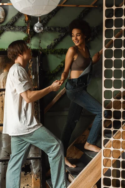 Sexy african american woman in overalls holding hands with boyfriend while walking upstairs in workshop — Stock Photo