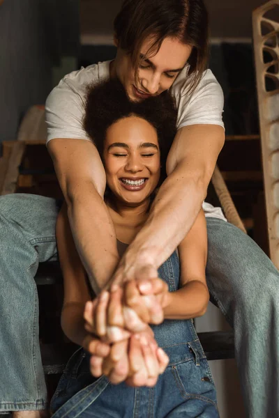 Uomo sorridente in jeans che si tiene per mano con allegra donna afroamericana seduta sulle scale con gli occhi chiusi — Foto stock