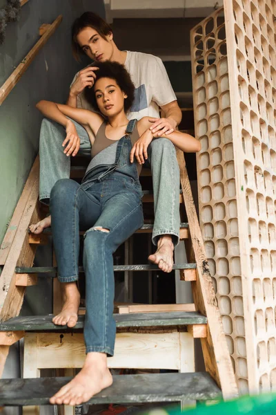 Young and barefoot interracial artists in denim clothes sitting on stairs in workshop and looking at camera — Stock Photo