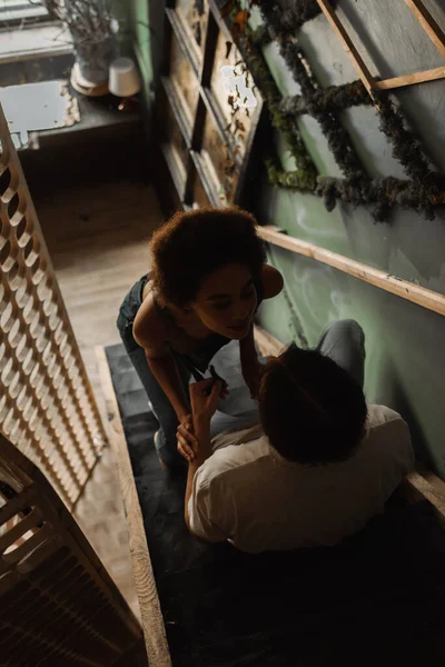 High angle view of sexy african american woman near young artist sitting on stairs in art studio — Stock Photo