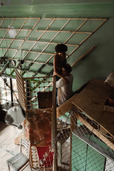 High angle view of young multiethnic couple embracing on stairs in workshop with decorative lattice — Stock Photo