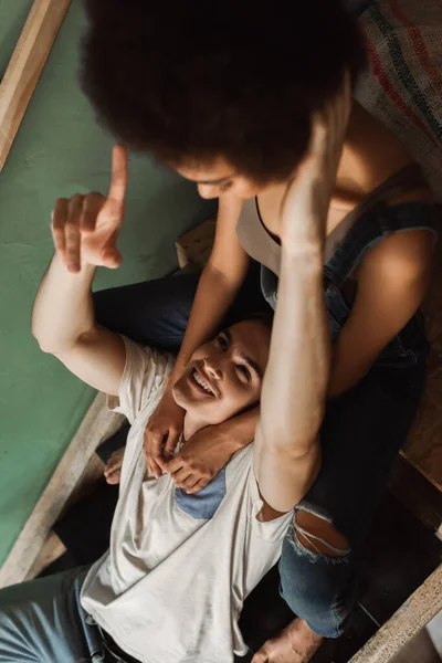 Vista de ángulo alto de hombre alegre señalando con el dedo cerca borrosa novia afroamericana en escaleras en el taller - foto de stock