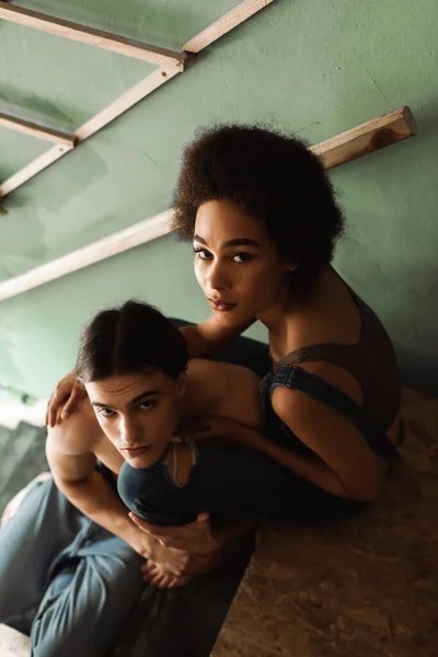 High angle view of young interracial artists embracing and looking at camera on stairs in art studio — Stock Photo