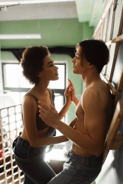 Vista lateral do jovem artista e mulher afro-americana em cima e macacão juntando as mãos e olhando uns para os outros na oficina — Fotografia de Stock