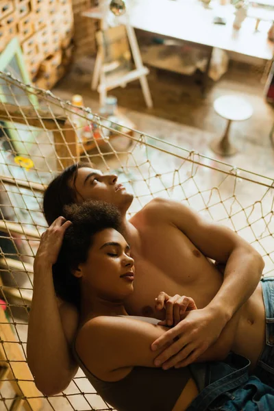 Homme torse nu rêveur étreignant jeune copine afro-américaine et regardant loin dans hamac en maille — Photo de stock