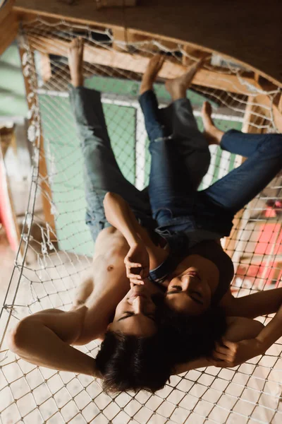 Vista superior de hombre sin camisa en pantalones vaqueros y mujer afroamericana en la parte superior y overoles acostado en hamaca de malla por encima del taller - foto de stock