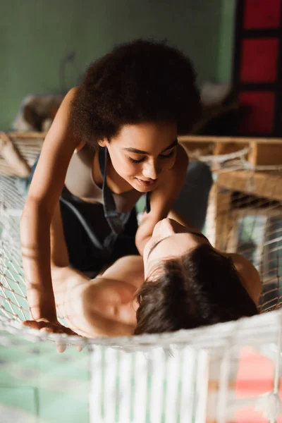 Passionate african american woman looking at shirtless man lying in net hammock in workshop — Stock Photo