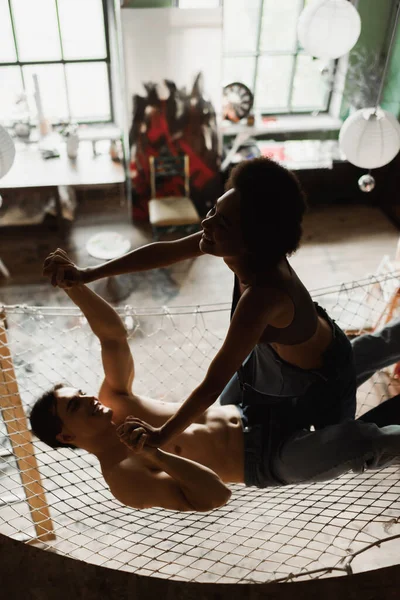 Веселый мужчина без рубашки, держась за руки с африканской американской подружкой, веселясь в гамаке над мастерской — стоковое фото