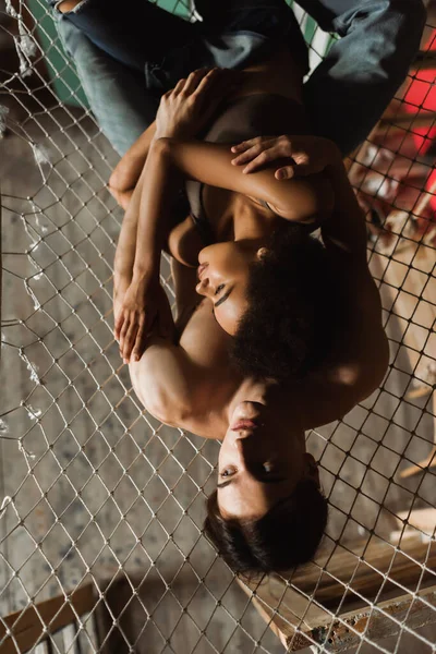 Top view of shirtless man and seductive african american woman embracing in hammock in art studio — Stock Photo