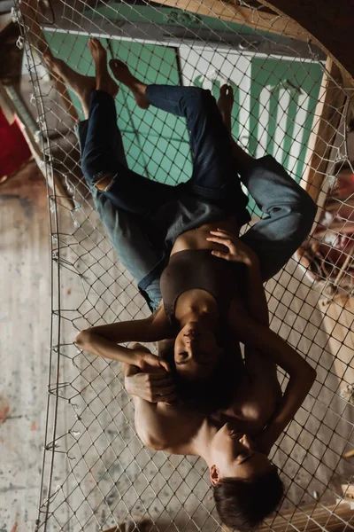 Вид зверху сексуальна афроамериканська жінка зверху і комбінезон, тримаючись за руки без сорочки в джинсах, лежачи в гамаку — стокове фото