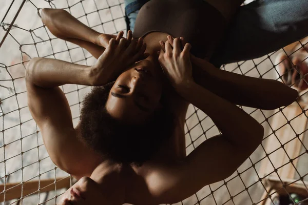 Top view of passionate interracial couple holding hands while lying in hammock in workshop — Stock Photo