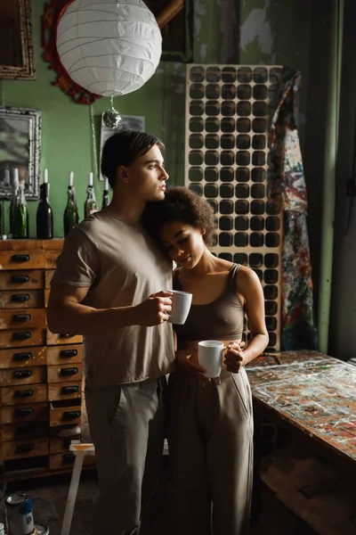 African american woman with cup of tea leaning on dreamy boyfriend looking away in workshop — Stock Photo