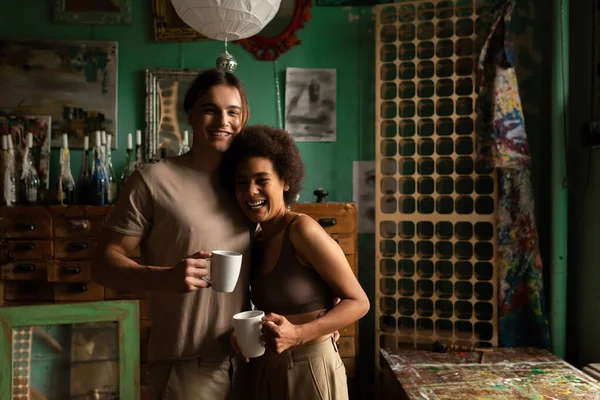Happy interracial couple with tea cups looking at camera in studio with vintage decor — Stock Photo