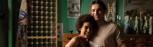 Young multiethnic artists looking at camera near bottles with candles in workshop, banner — Stock Photo