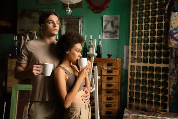 Young and dreamy artist looking away and hugging african american girlfriend drinking tea in workshop — Stock Photo