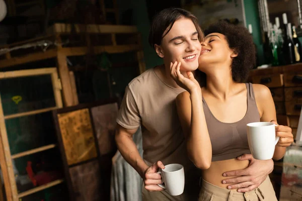 Seductora mujer afroamericana con taza de té besando hombre feliz en taller de arte - foto de stock