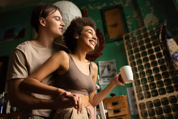 Low angle view of smiling artist holding hands with cheerful african american woman with tea cup in workshop — Stock Photo