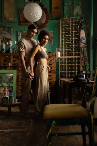 Sexy african american woman with cup of tea holding hands with boyfriend and smiling at camera in art studio — Stock Photo