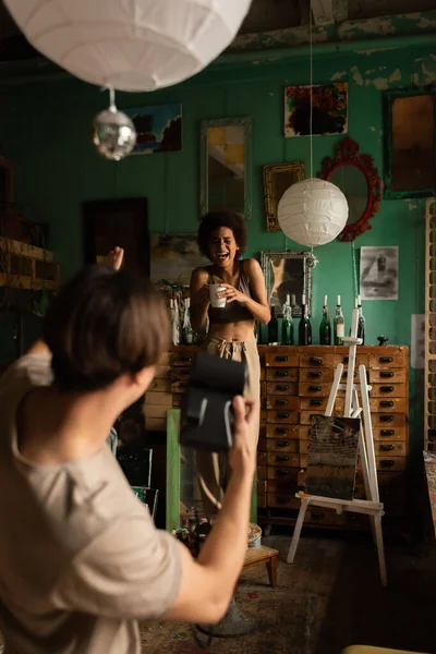 Alegre mujer afroamericana riendo y posando con taza de té cerca de hombre borroso con cámara vintage en estudio de arte - foto de stock