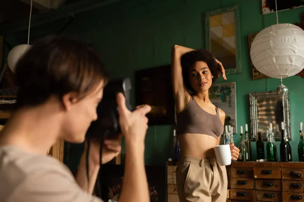 Sexy african american woman in top posing near blurred boyfriend with vintage camera — Stock Photo