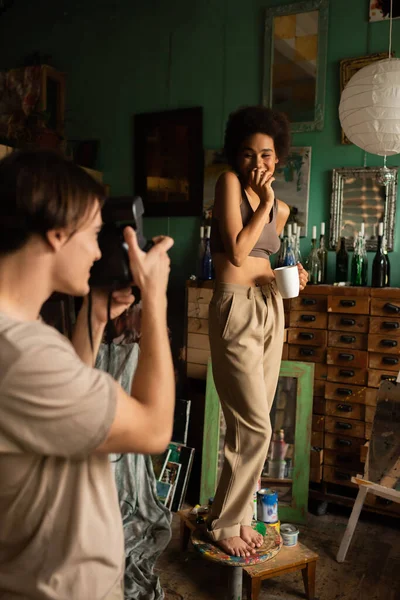 Homem desfocado com câmera vintage tirando foto da namorada americana africana em pé no banquinho com xícara de chá — Fotografia de Stock