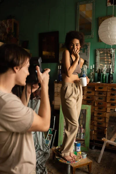 Homme flou avec caméra vintage prenant des photos de petite amie afro-américaine debout sur tabouret avec tasse de thé — Photo de stock