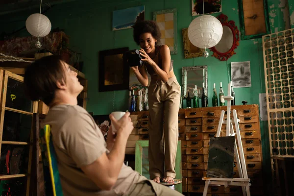 Cheerful african american woman taking photo of blurred boyfriend sitting with cup of tea in workshop — Stock Photo