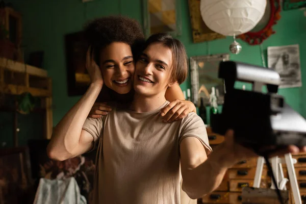 Jovem e feliz artista com câmera vintage borrada tirando foto de mulher americana africana alegre em oficina — Fotografia de Stock