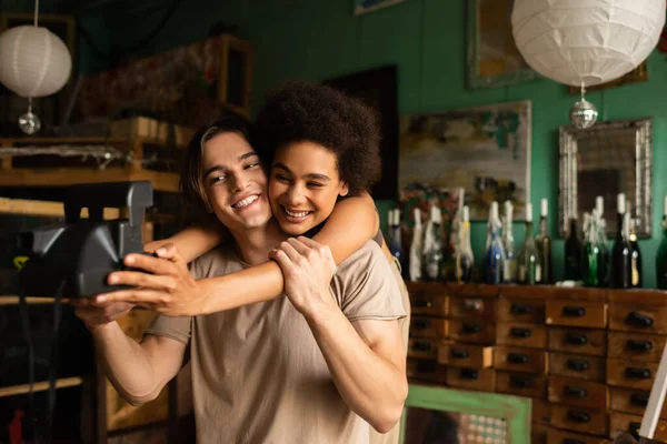 Bonita y feliz mujer afroamericana con cámara vintage tomando selfie con novio en taller con decoración vintage - foto de stock