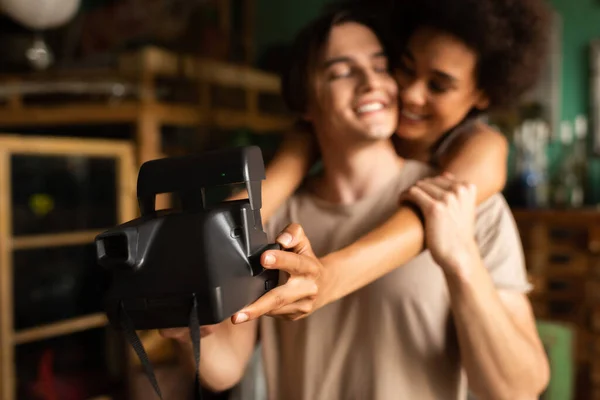 Blurred african american woman taking selfie on vintage camera with happy boyfriend in workshop — Stock Photo