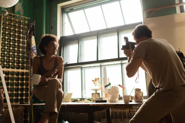 Hombre con cámara vintage tomando la foto de la sonriente mujer afroamericana sentada cerca de una gran ventana con taza y pinceles - foto de stock