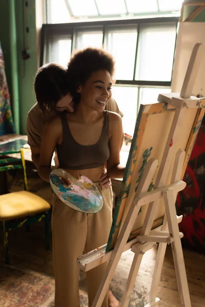 Young man kissing happy african american girlfriend standing with palette near easel — Stock Photo