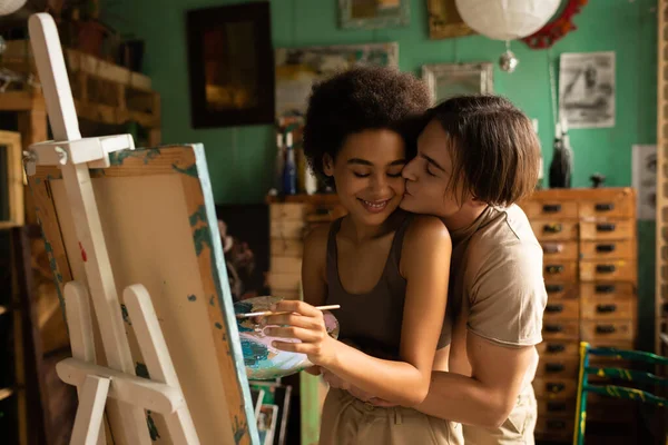 Happy african american woman painting on easel near young boyfriend kissing her in workshop — Stock Photo