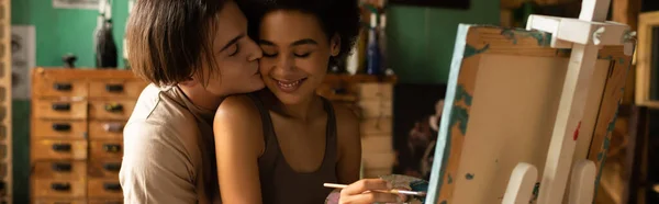 Young man kissing sensual african american woman painting on easel in workshop, banner — Stock Photo