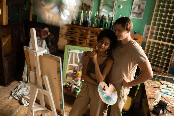 African american woman with palette and paintbrush looking at easel near man standing with hand on hip — Stock Photo