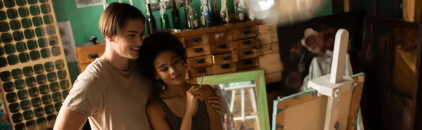 Happy interracial artists looking at easel in workshop, banner — Stock Photo
