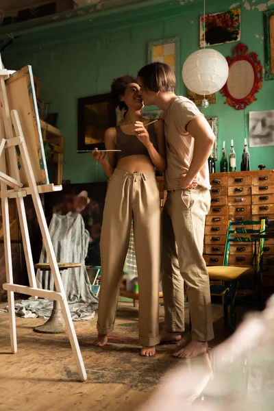 Pleine longueur de pieds nus interracial couple baisers près de chevalet dans l'atelier d'art avec décor vintage — Photo de stock