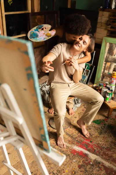 African american woman with palette and paintbrush kissing barefoot man pointing at easel while sitting in workshop — Stock Photo