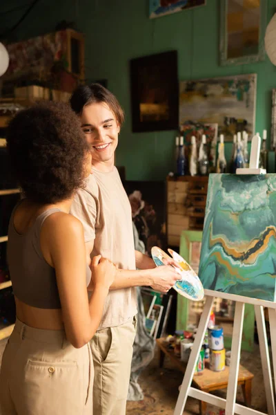 Heureux jeune artiste avec palette regardant afro-américaine copine près de peinture sur chevalet — Photo de stock