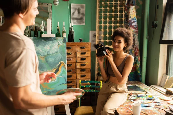 Souriant afro-américaine tenant caméra vintage près de petit ami debout avec palette près de chevalet — Photo de stock