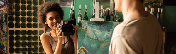 Happy african american woman with vintage camera taking photo of blurred boyfriend near painting on easel, banner — Stock Photo