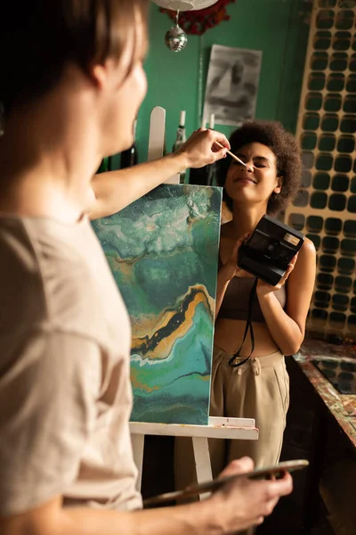 Blurred man painting on nose of cheerful african american girlfriend with vintage camera in art workshop — Stock Photo