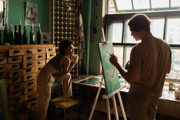 Jeune artiste peinture sur chevalet près séduisante femme afro-américaine avec tasse de thé — Photo de stock