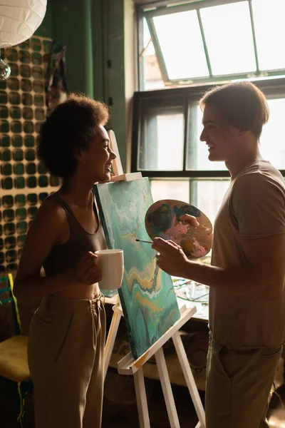 Side view of african american woman with tea cup and artist with palette smiling at each other near easel with painting — Stock Photo